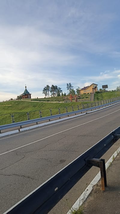 The image shows a view from a road in Kapyĺ, Belarus, looking towards a grassy hill with several structures. On the left side of the hill is a small wooden church with a green dome and a cross. Further to the right on the hill is a yellow building with a pitched roof and several windows. In between these two structures are several pine trees. In the foreground, a black metal guardrail with white markings runs along the side of the road. The road itself is asphalt with white lane markings. The sky is partly cloudy with a soft, diffused light, suggesting it might be late afternoon or early morning. The overall scene depicts a rural or semi-rural landscape with religious and possibly public buildings situated on a prominent hill overlooking the road.