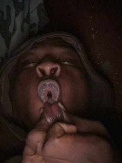 A close-up, low-light, overhead shot captures a person with dark skin in Ikorodu, Nigeria. Their eyes are closed, and their mouth is open, with a tongue visible inside. A finger is touching the tongue. The background is dark and out of focus, with some indistinct patterns that might be fabric.