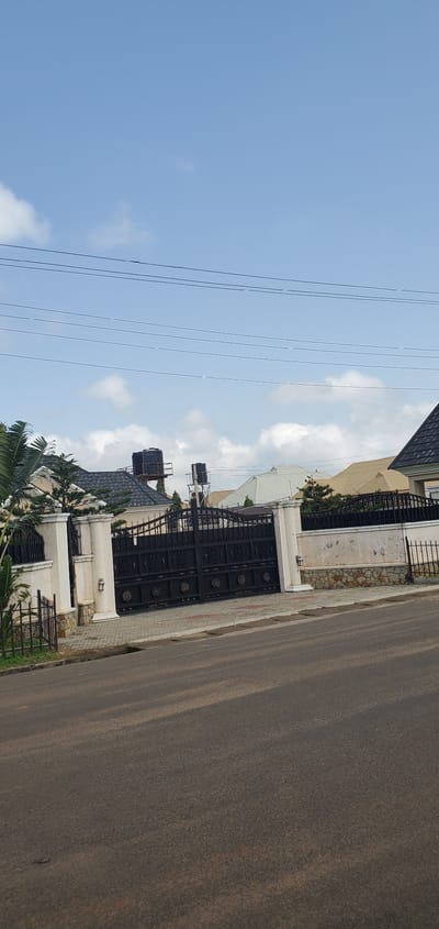 The image depicts a large, ornate black metal gate with intricate scrollwork, flanked by decorative white pillars and a stone-clad wall. Beyond the gate, the roofs of several houses are visible, some with dark grey shingles and others with lighter colored, possibly metal roofing. Two large black water tanks on elevated platforms are prominent atop one of the buildings. Trees and other vegetation are sparsely distributed around the properties. Overhead, several power lines are strung across a pale blue sky with scattered white clouds. The foreground shows a paved road with a distinct incline, suggesting a slight elevation change in the terrain. The setting appears to be a residential area, likely a well-to-do neighborhood given the substantial gate and property style. The scene is devoid of people or any activity, presenting a static view of the entrance to a private residence in Barakin Akawo, Nigeria.