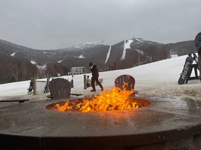 A fire pit burns brightly on a snowy mountain ski resort. Ski slopes stretch towards the horizon, marked by white snow and bare trees. A lone skier walks by the fire. Two Adirondack chairs sit near the fire, and snowboards rest nearby, indicating a place for relaxation after skiing. The cloudy sky suggests a cold, overcast day.