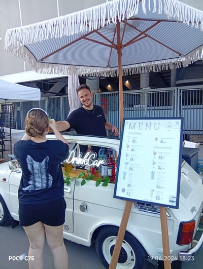 A man and a woman are standing by a white car converted into a bar. The car has a striped umbrella over it. The man is handing a drink to the woman. A menu board stands in front of the car. The scene appears to be outdoors, with a fence and stadium seating in the background. The text on the menu and the car indicate the location is "DrinkCar" and "Mojito." The date "19.06.2026" and time "18:23" are visible at the bottom right. The text on the woman's shirt is a black and white image of a cat.