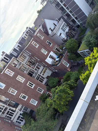 An aerial view, captured from a high vantage point, showcases a densely populated urban environment in Amsterdam, Netherlands, characterized by a mix of architectural styles and lush greenery. The image is tilted, creating a dynamic perspective of the cityscape. In the foreground, a light-colored, angular structure, possibly a balcony or rooftop edge, dominates the lower right portion of the frame. This structure casts a shadow that partially obscures the scene below.
Dominating the mid-ground is a large, brick-faced apartment building with numerous white-framed windows, some of which are open. The building's architecture suggests a historical style, typical of older European city centers. Adjacent to this brick building is a smaller, white multi-story building with balconies adorned with plants and outdoor furniture. This building appears to be connected to or integrated within the larger complex.
Further back, a modern, multi-story building with a facade of glass and dark panels stands out, offering a stark contrast to the older brick structure. Balconies with glass railings are visible on this building, hinting at contemporary apartment living.
The scene is interspersed with abundant trees and vegetation, creating courtyards and rooftop gardens. A notable feature is a small structure with a green, vegetated roof, likely a garden shed or a compact greenhouse, situated within a garden area. The garden contains various types of trees, bushes, and flowering plants, with a wooden table and chairs visible, suggesting an outdoor seating area.
The sky above is a pale blue with wispy white clouds, indicating fair weather. The overall impression is one of a vibrant, lived-in urban space where modern and older structures coexist, with a strong emphasis on green spaces integrated within the built environment. The angle of the photograph suggests it was taken from a higher building looking down and across at the adjacent structures and their internal courtyards. No people are clearly visible in the image.