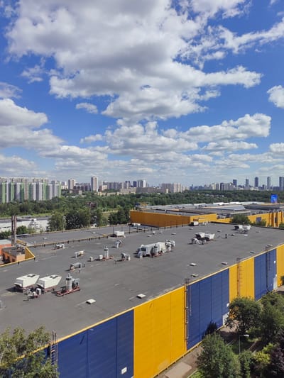 This aerial photograph, captured from a high vantage point, showcases a sprawling urban landscape beneath a partly cloudy blue sky. In the foreground, the flat, dark roof of a large industrial or commercial building dominates the scene, punctuated by numerous HVAC units and ventilation systems. The building's exterior features bold yellow and blue stripes along its side. Beyond this structure, a dense green forest stretches out, interspersed with trees. Further in the distance, a cityscape emerges, characterized by a cluster of high-rise residential buildings and taller modern skyscrapers. The photo is taken in Putilkovo, Russia. There are no people or animals visible in the image.