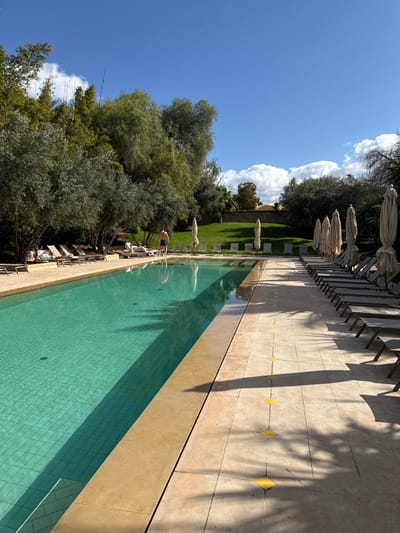 This image depicts an outdoor swimming pool area in Marrakesh, Morocco, under a bright blue sky with scattered white clouds. A long, rectangular swimming pool, tiled in teal, occupies the left side of the frame, with clear water reflecting sunlight and shadows from surrounding trees. The pool is bordered by light beige stone decking.
On the left side of the pool, a section of the deck is visible with several unoccupied beige lounge chairs. Further left, partially obscured by dense green foliage, including tall bamboo and olive trees, are at least two individuals. One person, wearing dark swimwear, stands near the edge of the pool, facing away from the camera. Another person is reclined on a lounge chair under the trees.
The right side of the frame features a wide expanse of the light beige stone pool deck. A long row of numerous, unoccupied wooden-framed lounge chairs with light-colored fabric is arranged facing the pool, extending into the background. Adjacent to these chairs is a line of large, closed, beige canvas umbrellas. Yellow, diamond-shaped markings are intermittently embedded in the stone deck, creating a visible path.
In the background, beyond the pool and deck, there are undulating green grassy mounds and additional trees under the clear sky. A light-colored building or wall structure is visible in the far background. Strong shadows are cast across the pool deck and into the water, indicating a bright, sunny day.