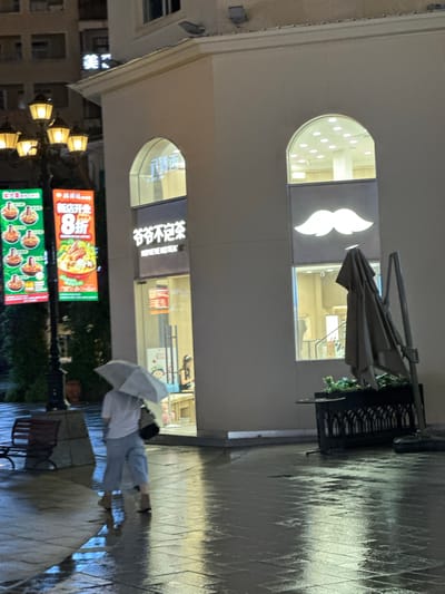 This image captures a nighttime street scene in the Chongqing High-tech Zone, China, following recent rainfall, evidenced by the wet, reflective tiled pavement.

The scene is dominated by the illuminated facade of a light-colored commercial building on the right, featuring two prominent arched windows. The ground floor houses a shop named "爷爷不泡茶" (Yéyé Bù Pàochá), with the English transliteration "ROYEYE NOTEA" visible below. To the right of this entrance, a large, glowing white mustache logo is displayed, partially in front of a lower arched window. An upper arched window reveals an interior with numerous recessed circular lights on the ceiling.

On the left side of the frame, a multi-lamp streetlight with five glowing yellow fixtures stands next to two large, vertical LED billboards. The left billboard, with a green background, displays multiple images of noodle bowls. The right billboard, with an orange-red background, announces "新店开业" (New Store Opening) and "8折" (20% off/80% discount), featuring a large image of a seafood noodle dish and the English text "SHRIMP SHRIMP SHRIMP." In the background, a dimly lit building shows a partial neon sign displaying the Chinese character "美" (Měi).

A single individual walks from left to right, their back to the viewer, holding an open light-colored umbrella. They wear a white short-sleeved shirt, light blue/grey loose-fitting pants, and dark flat shoes, carrying a dark shoulder bag. Their reflection is visible on the wet pavement.

In the foreground to the right, a dark decorative planter box with artificial greenery holds a folded grey/brown outdoor umbrella. A dark park bench is partially visible on the far left, behind the streetlight. The overall lighting is artificial, emanating from the streetlights, billboards, and building interiors, creating strong reflections on the glistening wet ground.