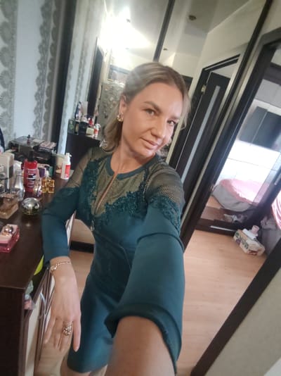 A woman is taking a selfie in an indoor setting. She has blonde hair and is wearing a dark teal dress with lace detailing on the shoulders and neckline. She is also wearing jewelry, including earrings, a bracelet, and a ring. The background appears to be a room with a dresser or vanity to her left, filled with various bottles and containers. To her right, there is a large mirrored wardrobe reflecting a bed and a box on the floor. The flooring is light-colored wood. There is no visible indication of the city of Petropavlovsk-Kamchatsky, Russia.