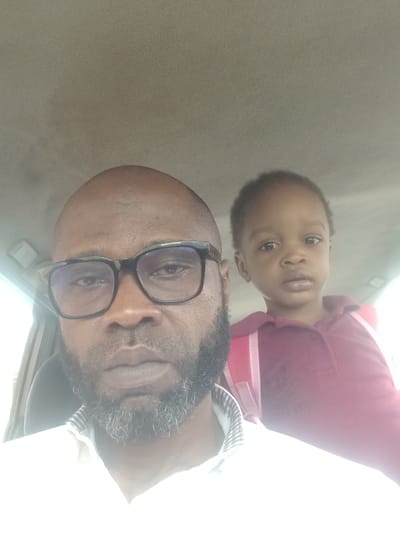 The image features an adult male and a young child, likely a father and child, captured in an indoor setting in Barden Yana, Nigeria. The photograph is taken from a slightly low angle, with the adult male in the foreground, and the child positioned behind and slightly above him. The adult male has a bald head and a full, dark beard with some visible grey strands. He wears dark-rimmed rectangular glasses, through which his eyes are visible, and appears to be wearing a collared shirt with a striped pattern on the cuff or collar area.

The child, appearing to be a toddler or young pre-schooler, has short dark hair and a light brown complexion. They are wearing a red or maroon polo shirt with some light-colored striping visible on the side. The child looks directly at the camera with a neutral, slightly curious expression and wide eyes. Both subjects have neutral emotions.

The background is a uniform, plain light gray or beige color, suggesting a simple wall or interior of a vehicle. A strong light source, possibly sunlight, enters from the right side of the frame, causing significant overexposure and glare, particularly on the right side of the adult's face, shirt, and parts of the background, indicating it is likely daytime. No text is visible in the image.