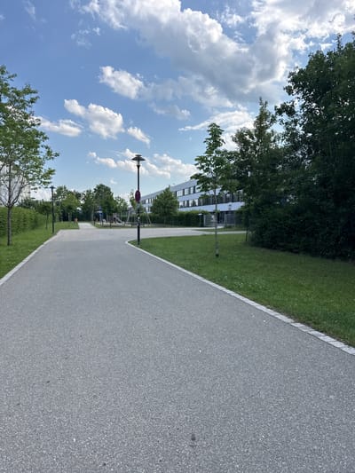 This image depicts an outdoor scene in Höhenkirchen-Siegertsbrunn, Germany. The perspective is from a paved pathway, suggesting a park, recreational area, or a path alongside a building. The pathway is a light gray asphalt with a white border line, running diagonally across the frame from the bottom right to the upper left. To the left of the pathway, there is a well-maintained lawn with scattered trees and bushes.

In the background, a modern building with a white facade and numerous windows is visible. There are also children's playground elements, including swings and a slide, interspersed with trees. Streetlights are positioned along the pathway, with one prominent lamp with a circular fixture visible closer to the foreground. A red and white circular sign with a diagonal bar, often indicating a prohibition, is mounted on a lamppost.

The sky is a bright blue, dotted with fluffy white clouds, suggesting a pleasant, partly cloudy day. The presence of lush green grass and trees indicates that it is likely daytime, possibly late morning or early afternoon, and the weather is fair. There are no people visible in the image, and the overall mood is peaceful and serene. The scene is characteristic of a public outdoor space designed for recreation and walking.