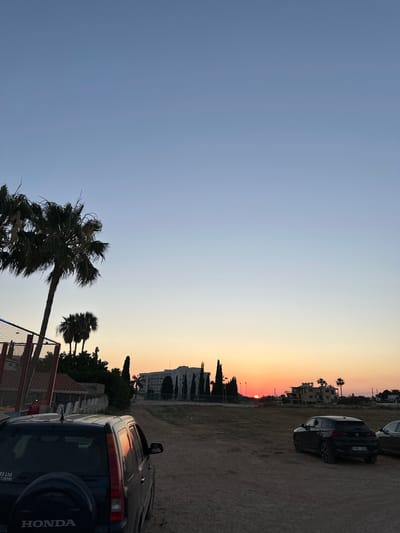 The image captures a sunset scene in the city of Peyia, Cyprus. In the foreground, the rear of a dark-colored Honda CR-V is visible, parked on a dirt road. To the right of the Honda, a black BMW X2 is parked. In the background, a large white building complex stands amidst sparse vegetation, with palm trees and other trees scattered throughout the landscape. The sun is setting on the horizon, casting an orange glow across the sky and reflecting on the side of the Honda. The sky transitions from a light blue at the top to shades of orange and yellow near the horizon. The environment appears to be a semi-arid or undeveloped area on the outskirts of a town.