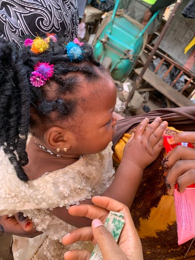 A child lies on their back, adorned with colorful flower hair accessories and earrings. An adult's hand holds currency, and another's fingers clutch a pink package with text visible. The child wears a cream-colored, floral-patterned garment, and the surrounding textiles display intricate designs. A teal chair is visible in the background. The setting appears cluttered.