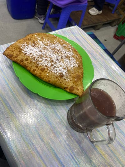 A fried turnover dusted with powdered sugar rests on a bright green plate, alongside a large glass mug filled with a dark, murky beverage. Both sit on a table with a light-colored, textured surface. Hints of chairs, including a bright blue one, are visible in the background. The composition is simple, showcasing the food and drink as the primary subjects.