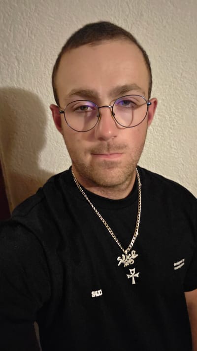 A close-up, waist-up, indoor portrait shows a young man with short, dark hair and light skin wearing a black t-shirt, glasses, and a silver chain with a pendant. The background is a textured wall, casting a shadow on the left. The man is looking directly at the camera with a neutral expression. There is text on his t-shirt, with "SALO" visible on the left side and "designed by moneyp***" on the right. The man appears to be indoors, possibly in a residential setting. There are no specific indicators suggesting this location is in Bania, Bulgaria.