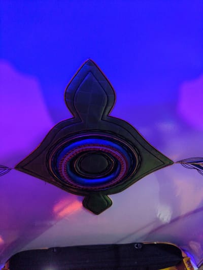The image features an intricately designed, stylized object displayed against a dramatically lit background. The primary subject is a dark, possibly leather or padded fabric, object shaped like an elongated diamond or a futuristic shield, with pointed ends along its horizontal axis. It is bordered by a metallic gold trim, and its surface exhibits a subtle quilted or stitched pattern.

At the center of this object, several concentric rings are visible, glowing with vibrant blue and purple light, suggesting internal illumination or highly reflective material under colored lights. Some inner rings also show a reddish-brown textured pattern. Thin black strings or wires extend from the top and bottom points of the object, adding to its visual complexity.

The background lighting is striking, with the left side of the wall bathed in a strong, vivid blue. This color transitions smoothly into purple towards the center and right side of the image. The far right portion of the background appears lighter, possibly white, reflecting the purple and a soft orange-red glow. On the extreme right, a dark, indistinct structure is partially visible, featuring a small, circular metallic emblem that resembles a stylized lowercase 'e'.

The overall scene suggests an artistic installation or a decorative display, likely in a modern exhibition space, a conference venue, or a contemporary setting, possibly in Bengaluru, India. The lighting creates a captivating and somewhat mysterious atmosphere. There are no people visible, and no specific activity is taking place other than the presentation of this object as a visual piece.