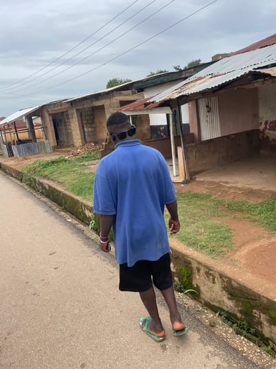 A man is walking on a paved road next to a grassy ditch and buildings. He is wearing a blue t-shirt, black shorts, and green flip-flops. He has sunglasses on and is holding a black smartphone in his right hand. The buildings appear to be made of brick and corrugated metal, with some showing signs of disrepair. There are power lines visible in the sky, which is overcast, suggesting it might be a cloudy day. The scene appears to be a street in a rural or semi-urban area in Barakin Lerawi, Nigeria. The man's posture suggests he is walking casually, and there are no strong emotional cues visible.