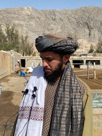 A man with a dark beard and turban stands outdoors. He is wearing a brown-patterned shirt, a shawl with gray and tan stripes, and a white embroidered cloth is draped over his shoulder, bearing three lapel microphones. In the background is a mountainous terrain, and what appears to be a small, rural structure. A man is visible in the background wearing a white robe and a blue container. The scene is in a rural, possibly agricultural setting. There is no clear evidence to confirm this photo was taken in Kandahar, Afghanistan.