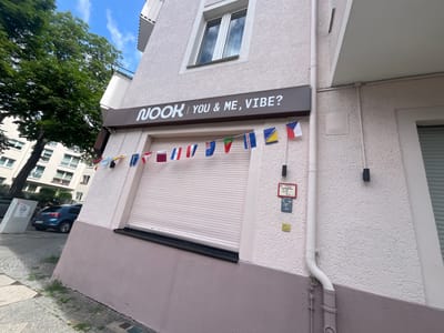 A low-angle, eye-level outdoor shot captures the exterior of a commercial building in Berlin, Germany, on a bright, cloudy day. The building's facade is textured and painted in a pale pinkish-gray hue, with a darker pink base. A white downspout runs vertically along the wall. Above the downspout, there are two red and blue rectangular signs with white text, possibly indicating building or utility information. A string of small, colorful international flags hangs across the front of the building above a closed, light-colored rolling security shutter. Above the shutter, a dark brown sign with white lettering reads "NOOK | YOU & ME, VIBE?". A window with a white frame is visible on the upper floor. To the left of the building, a large green tree with dense foliage stands tall, and part of a multi-story residential building with balconies and windows is visible behind it. On the street in front of the commercial building, a blue hatchback car is parked. The sidewalk is paved with cobblestones and concrete slabs, with patches of green grass growing between them.