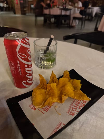 The image shows a table at a Chinese restaurant, likely in Kuta, Indonesia, with a meal consisting of fried wontons, a can of Coca-Cola, and a glass of water with a straw. The wontons are golden brown and arranged on a black rectangular plate lined with paper that has red Chinese characters and English text that reads "PANDA CHINESE RESTAURANT" and "OPEN LUNCH AND DINNER DAILY 11AM - LATE". The Coca-Cola can is red with the iconic white script logo and the text "FIFA WORLD CUP™ OFFICIAL PARTNER". The can is covered in condensation, suggesting it is cold, and the glass of water also has condensation and contains ice and what appears to be lime slices, with a black straw. The table is white, and the background is blurred, showing what appears to be other diners and restaurant furniture, indicating an indoor setting during daylight or under artificial light. The scene evokes a casual dining experience, possibly a lunch or dinner outing.