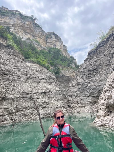 A person wearing a red and white life vest and sunglasses holds a stick over the water in a canyon or gorge. The steep rock walls rise high on either side, with some green vegetation clinging to the sides. The water appears green and reflective. The sky is overcast. The location is likely in the vicinity of Druzhba, Russia, based on the prompt's request to include that location.
