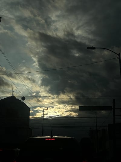 The image depicts a street scene in Puebla, Mexico, during what appears to be either dawn or dusk. The sky is dramatically overcast with dark, heavy clouds interspersed with patches of brighter light breaking through, suggesting either the sun is rising or setting. Silhouetted against the sky are several urban elements: a building with an antenna on its roof is visible on the left, and a street light with a curved arm extends from the right side of the frame. Numerous electrical wires crisscross the scene horizontally. In the foreground, the dark silhouette of a vehicle's roof is visible, with its brake lights illuminated. The overall impression is one of a subdued, potentially atmospheric urban environment.