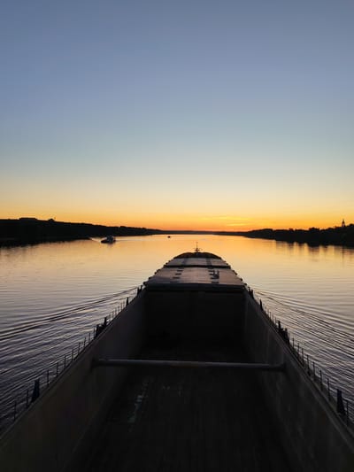 The image depicts a wide, calm river at sunset, viewed from the stern of a large barge. The sky transitions from a pale blue at the top to a vibrant orange and yellow near the horizon, casting a warm glow on the water. The water's surface is rippled, indicating the barge's movement.

Along the riverbanks, silhouetted trees and buildings form a dark, elongated line. In the distance, on the left side of the river, a small white boat is visible, moving away from the viewer. On the right bank, a prominent domed structure, possibly a church, stands out against the darkening landscape. The barge itself is a dark, imposing presence in the foreground, its open cargo hold visible. The scene conveys a sense of tranquility and the vastness of the river.

Based on the presence of the domed structure often associated with the city's historical architecture and the wide Volga River, this scene is likely located in Yaroslavl, Russia, possibly along the Volga River embankment near the assumption cathedral.