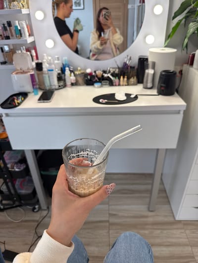 A person's hand holds a glass of iced coffee with a clear plastic straw. The background features a vanity table with various makeup products, brushes, and a circular mirror with lights. The vanity is set up in what appears to be a home or studio environment with wooden flooring and a white drawer unit. The reflection in the mirror shows two women, one of whom is holding a makeup brush and appears to be interacting with the other, who is taking a photo with her phone. The overall impression is a behind-the-scenes glimpse into a makeup or beauty routine. There is no information in the image to indicate the location is in Riga, Latvia.