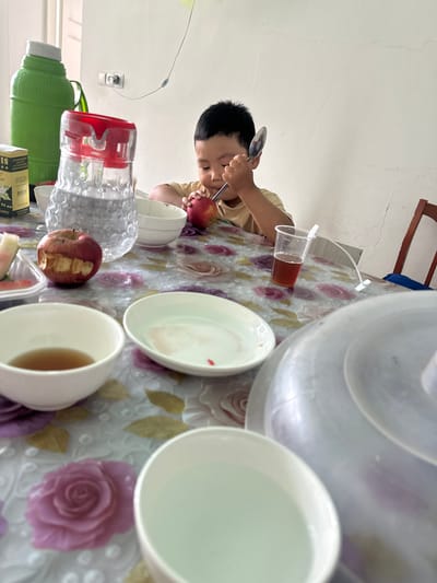 A young boy is sitting at a table in a room with a light-colored wall, possibly in a residential setting in Тайгараев, Kyrgyzstan. He is wearing a light-colored shirt and holding a spoon, engaged in eating an apple. On the table, there are several bowls, some with food or liquid, a pitcher of water, a plastic cup with a drink, and a large green thermos. The table is covered with a floral-patterned tablecloth. The overall scene depicts a mealtime in progress.