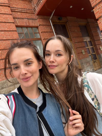 Two young women are taking a selfie outdoors in front of a brick building. The woman on the left is closer to the camera and is looking directly at it with a slight smile. She has brown hair pulled back in a bun and is wearing a denim jacket over a grey shirt. The woman on the right is slightly out of focus and is looking to her right. She has long brown hair and is wearing a white shirt with a colorful patterned strap over her shoulder. The building in the background is made of red brick with large windows. The context of the image is the facade of the Bashkir State Academic Drama Theater named after M. Karim in Ufa, Russia, which is visible with its architectural details.