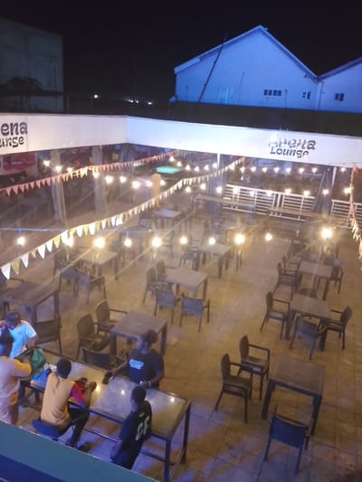 The image displays an outdoor lounge area identified by signage as "Athena Lounge," located in Narayi, Nigeria, at night. The scene is illuminated by numerous overhead string lights featuring exposed bulbs and triangular bunting.
The primary area consists of multiple dark-colored square and rectangular tables, predominantly unoccupied, each accompanied by several matching chairs, arranged on a light-colored paved surface.
Six individuals are visible. In the lower-left foreground, four persons are clustered around a rectangular table with a glass top. One individual in a light-colored shirt is standing, leaning over the table. Another, in an orange short-sleeved top, stands adjacent. A third, wearing a yellow shirt, is seated on a dark stool, engaged with a mobile device. A fourth, wearing a dark t-shirt with "CF" on the back, is also seated at this table. Towards the center-left, a person with dark, curly hair, wearing a black t-shirt with "Athena Lounge" printed on it, stands near an empty table, holding a phone. A sixth individual is partially visible standing near a green-felt pool table in the upper-left midground.
In the background, under a dark night sky, prominent white buildings with pitched roofs are visible. White railings demarcate elevated sections of the lounge.
