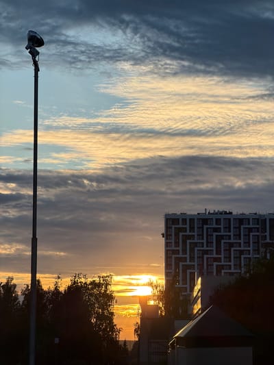 The image depicts a dramatic sunset over an urban landscape, likely in Nizhnekamsk, Russia. The sky is filled with a striking contrast of dark, streaky clouds on the left and bright, golden clouds illuminated by the setting sun on the right. The sun itself is a brilliant orb peeking through the horizon, casting a warm glow that reflects off a modern, multi-story building with a distinctive geometric facade.
In the foreground, a tall lamppost with a single, dark lamphead is positioned vertically, its stark silhouette cutting across the colorful sky. Trees, silhouetted against the bright sunset, frame the right side of the scene, adding a natural element to the urban setting. The building appears to be residential or commercial, with numerous windows visible.
The time of day is clearly sunset, indicated by the low angle of the sun and the vibrant, warm colors in the sky. The weather appears to be clear with scattered clouds, creating a visually dynamic atmosphere. The overall mood is one of awe and beauty, with the contrasting elements of the natural sky and the man-made structures creating a striking composition. No visible text is present in the image.