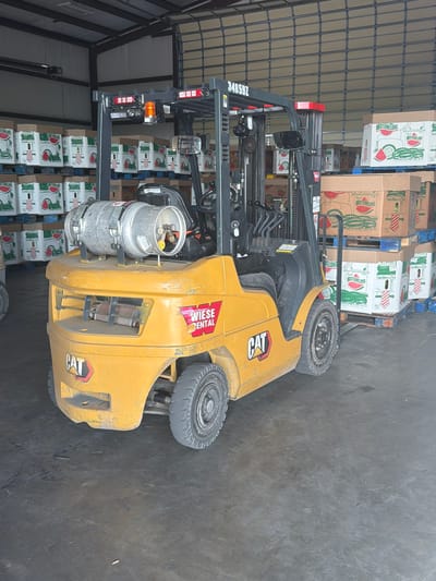 A yellow Caterpillar forklift is parked indoors in a warehouse setting in Hornersville, United States. The forklift is positioned in front of stacks of cardboard boxes filled with watermelons, some of which are labeled "Fresh Watermelons." The warehouse has a concrete floor and a large overhead garage door in the background. The forklift is equipped with a propane tank mounted on the back. The overall environment suggests a distribution or storage facility for produce. There are no people visible in the image.