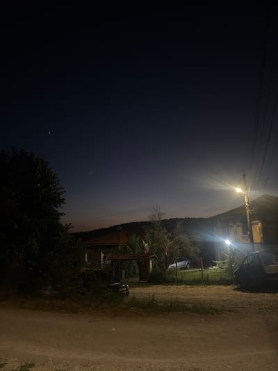 A nighttime scene depicts a rural setting, likely in Zlataritsa, Bulgaria. The sky is a deep indigo, transitioning to a faint orange and pink hue near the horizon, suggesting either dawn or dusk. Three celestial bodies, possibly stars or planets, are visible in the upper portion of the sky.

In the foreground, a dirt road leads towards a residential area. On the right side of the image, a building with a tiled roof is illuminated by a bright street lamp mounted on a wooden pole, casting lens flares. Another, smaller light source illuminates the front of the building. A dark-colored utility vehicle is parked to the right of the building.

To the left of the road, a dense tree with dark foliage occupies the left edge of the frame. Further into the scene, several other buildings are visible, including one with a red-tiled roof and a covered entryway. A light-colored car is parked in front of this building. Another dark-colored car is partially obscured by vegetation. A small structure with a flag on top is also present. The overall impression is of a quiet, dimly lit village at night.