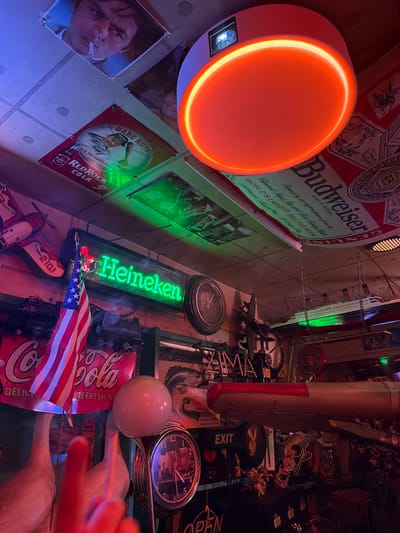 This media file depicts a dimly lit, eclectic interior, possibly a bar or man cave, adorned with a variety of memorabilia. Dominating the upper portion is a large Budweiser advertisement. To the left, a circular orange light fixture casts a warm glow. Scattered throughout the scene are vintage posters featuring Babe Ruth and Red Rock Cola, along with a neon Heineken sign. Various objects are arranged, including clocks, model airplanes, and what appears to be a propeller from a vintage aircraft. A prominently displayed American flag adds a patriotic touch. There are no people clearly visible, but a portion of a person's hand is visible in the lower right corner, and a cigarette is visible in the mouth of a person depicted in a poster on the left. The overall ambiance suggests a nostalgic and perhaps masculine space, filled with collected items from different eras and brands. The lighting is moody, with strong contrasts between illuminated areas and shadows, creating a cozy and perhaps slightly cluttered atmosphere. The presence of vintage advertisements and collectibles points to a possible theme of Americana or retro branding. The location context of Adachi, Japan, is not visually evident from the image itself, but it is provided as additional information.