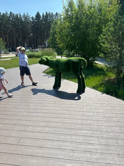 On a sunny day, two children and a topiary sculpture of a lamb are in a park. A boy, wearing a white t-shirt with a pixelated pattern, dark shorts, a light green bucket hat, and sandals, stands on a wooden deck, his right hand touching the brim of his hat. To his left, a young girl in a white t-shirt, pink shorts, a light blue hat, and sandals walks across the deck. In the middle ground, a green topiary sculpture of a lamb stands on the grass. Behind the children and the sculpture, a line of tall pine trees forms the background, with a sign that reads "НАШЕ КАФЕ" (Our Cafe) visible amongst the foliage. The environment includes a wooden boardwalk, grass, and trees under a clear blue sky.