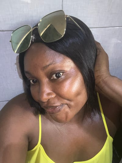 An adult female is depicted in a close-up, head-and-shoulders shot. She has dark skin, short, straight dark hair, and is wearing a yellow spaghetti strap top. Large, square-framed sunglasses with gold-colored frames and light-colored lenses are positioned on top of her head. Her left hand is raised, with fingers touching the side of her head. She is looking directly at the camera with her lips slightly parted. The background consists of a light-colored, possibly tiled or textured wall with horizontal lines. This scene is located in the city of Dzhidu, Nigeria.