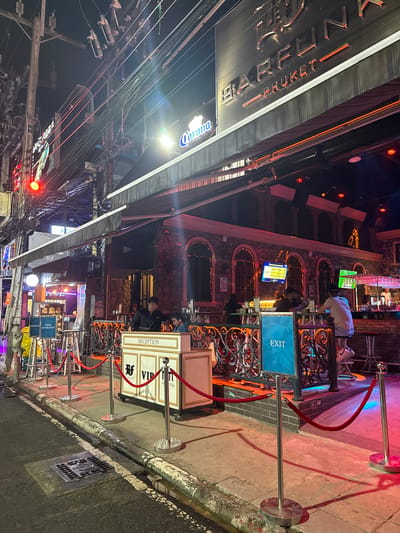 The image shows the exterior of a bar called "BARFUN" in Patong, Thailand, at night. The bar has a prominent sign with neon lights. The entrance is cordoned off with red velvet ropes and stanchions, and there is a reception desk. People can be seen inside the bar, some sitting at the bar, and others standing. The street in front of the bar is wet, suggesting it may have rained recently. There are also street lights and utility poles with many wires visible. A traffic light is illuminated red. The overall atmosphere is vibrant and lively, indicative of a nightlife scene.