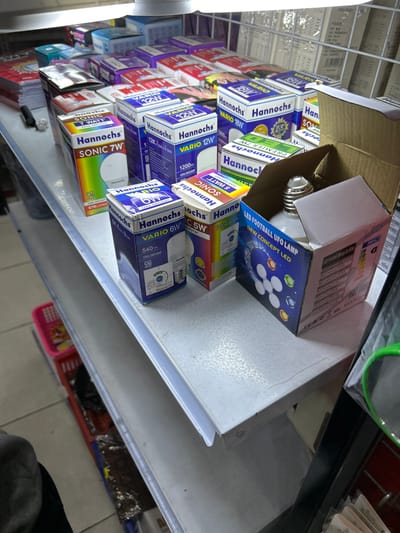 The image shows a retail display of LED light bulbs in a store in Depok, Indonesia. The shelves are stocked with various brands and types of bulbs, including Hannochs Sonic 7W, Hannochs Vario 12W, Hannochs Vario 18W, Hannochs Vario 6W, and Hannochs Vario 5W. One box is open and displays a "LED FOOTBALL UFO LAMP NEW CONCEPT LED" bulb. The lighting is bright and consistent, typical of an indoor retail environment. The overall scene is one of product display and organization.