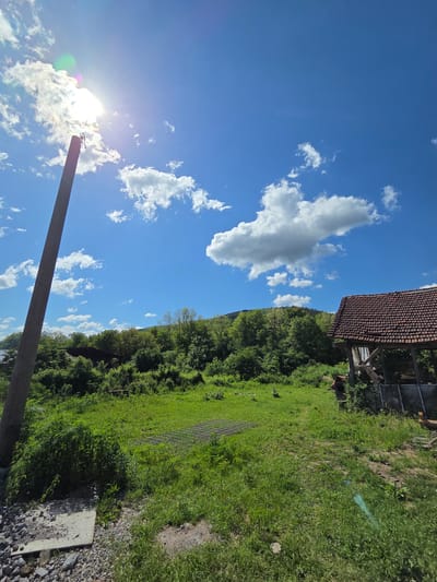 The image depicts a rural scene in or near Kaleytsa, Bulgaria, under a partly cloudy sky. In the foreground, a weathered wooden pole is visible, positioned against the bright sun. In the middle ground, a grassy field slopes upwards, bordered by a dense line of trees. A wire fence runs along a portion of the field. An aged, partially dilapidated structure with a tiled roof is visible at the top of the slope. The sky is blue with scattered white clouds.