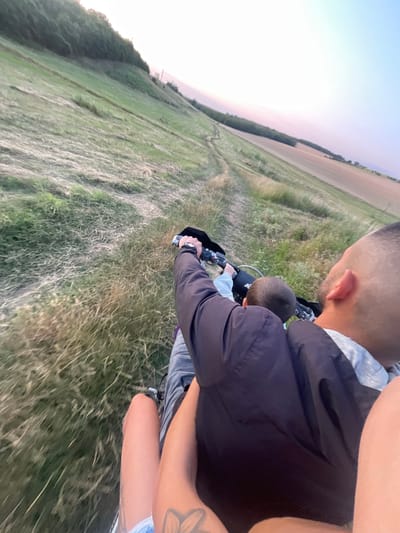 The image depicts a first-person perspective of two individuals, an adult and a child, riding on a dirt bike through a grassy, hilly terrain. The adult, facing away from the camera, is operating the dirt bike, with their left arm visible on the handlebars. The child is positioned in front of the adult, also facing away from the camera, with their head visible. The environment is characterized by a steep, grassy slope with patches of dry grass and a dirt path winding upwards. In the background, a line of trees separates the grassy slope from a cultivated field under a hazy sky with subtle pink and blue hues, suggestive of dusk. The overall scene is rural and appears to be located in Zheglartsi, Bulgaria.