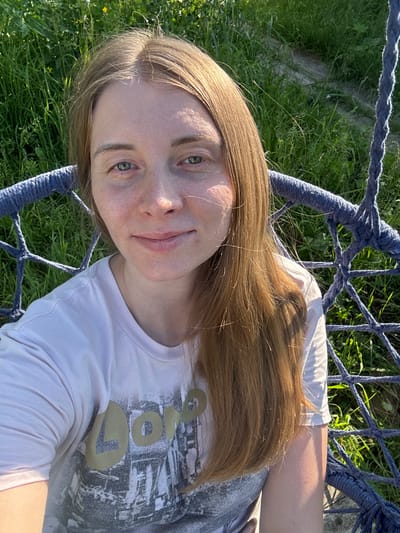 A young woman with long, auburn hair is sitting in a blue rope swing outdoors, with a background of green grass and vegetation. She is wearing a white t-shirt with a graphic print. Her head is tilted slightly, and she is looking towards the camera with a subtle smile. The lighting suggests it is daytime, with sunlight casting shadows on her face and clothing. The scene appears to be in a natural outdoor setting, possibly a park or a backyard. There is no indication of the city of Kurgan, Russia.