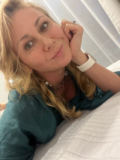 A woman with blonde hair and fair skin is looking at the camera. She is wearing a dark green top and a white watch. She has on a silver necklace. Her head is resting on her hand, which has a ring on the ring finger. A white textured fabric is in the background. The location cannot be determined.