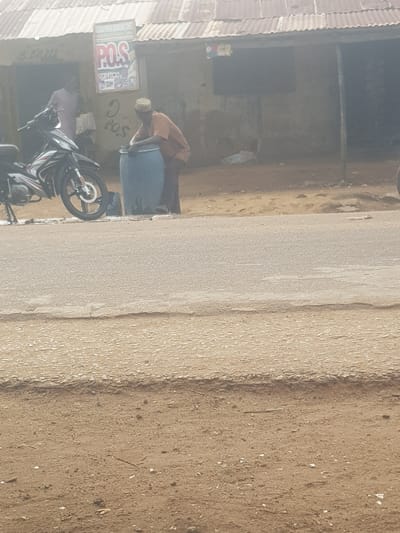 The image displays an outdoor street scene in a semi-urban or rural setting, likely Kufena, Nigeria, under bright daytime conditions. A paved road runs horizontally across the middle ground, bordered on the right by a wide, light brown dirt or sand shoulder. On the left side of the road, there is a building with a corrugated metal roof and light brown, textured walls.
In front of this building, a man wearing an orange or brown shirt and a white skullcap (kufi) is bent over a large blue plastic barrel, appearing to be engaged in an activity like filling or dispensing. Below the barrel and slightly to its left, a black and white motorcycle with visible disc brakes is parked. Attached to the building's wall, above the man, is a rectangular sign prominently displaying "POS SERVICE." Below this sign, the word "POS" is also scrawled directly onto the wall. A second person is partially obscured behind the parked motorcycle.
The scene depicts a typical roadside environment, possibly a local shop or service point, with bright, even lighting consistent with a sunny day. No strong shadows are cast, indicating either high sun or diffuse light.