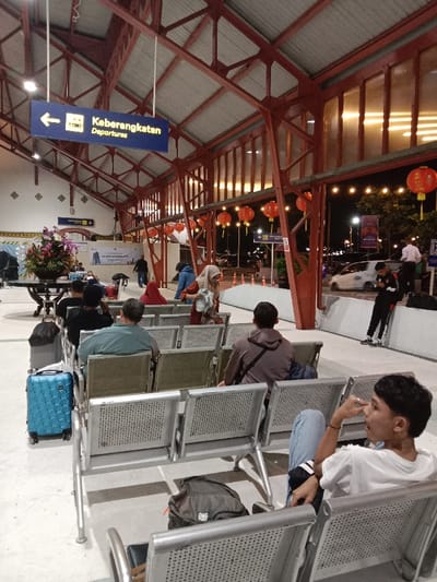 The image displays a nighttime scene within the semi-open departure hall of a train station in Semarang, Indonesia, identifiable by the architecture and "Keberangkatan Departures" signage, likely Stasiun Semarang Tawang. The hall features a distinctive red metal truss roof structure supporting a white corrugated ceiling, illuminated by overhead lighting. Numerous red spherical Chinese lanterns and string lights are strung along the right side of the trusses, extending into the background.

A blue overhead sign with a white arrow and a train icon reads "Keberangkatan Departures." In the foreground, a young male with short dark hair, wearing a white t-shirt and light blue jeans, is seated on a silver perforated metal bench, his right hand near his mouth. A dark grey backpack rests on the adjacent seat. Further back and to the left, a bright blue hardshell rolling suitcase stands beside other seated passengers, including one in a brown hooded sweatshirt and another in a teal shirt, all facing away from the camera.

In the midground, a woman wearing a light brown hijab and patterned top is standing near seating, holding a white item. Other figures are visible in the background, some walking. On the far left, a large floral arrangement containing purple and pink flowers is displayed on a dark circular table, with a white banner behind it. Outside the covered departure area on the right, a young male in a black and red jacket and dark pants leans against a low concrete wall, focused on a mobile phone. Vehicles, including a white car, are visible on a street beyond the station, illuminated by streetlights and other ambient light sources.