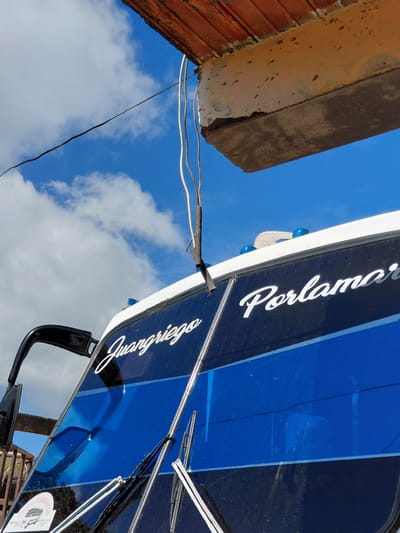 The image captures an outdoor scene, likely in Santa Ana, Venezuela. A dark blue bus with white lettering "Juangriego" and "Porlamar" is prominently featured. The bus has a black side mirror and windshield wipers. Above the bus, a section of a building with red tile roofing and a concrete overhang is visible against a blue sky with white clouds. Wires are hanging down from the overhang, partially obscuring the bus and the sky. The overall impression is of a somewhat weathered environment, possibly a bus terminal or a roadside view.