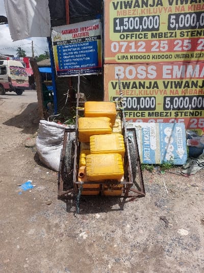 The image depicts an outdoor scene in Dar es-Salaam, Tanzania, specifically referencing Mbezi and Kiluvya areas based on prominent signage. In the foreground, a two-wheeled, unpainted metal handcart is positioned on dry, uneven dirt and gravel ground. The handcart carries a stack of at least five rectangular yellow plastic jerrycans, some secured with blue cordage. To the left of the handcart, a large, partially filled white woven sack rests on the ground.
Dominating the mid-ground are several large advertising banners. On the left, a white banner with blue text advertises "OKOMBOZI EDUCATION INSTITUTE," listing courses such as "COMPUTER COURSE," "SECRETARIAL COURSE," "GRAPHIC DESIGN," and "VIDEO PRODUCTION." To its right, two large yellow and green banners, stacked vertically, advertise "VIVANJA" (plots of land) in "MBEZI, KILUVYA NA KIBAHA," citing prices ranging from "1,500,000 - 5,000,000" and providing a contact number "0712 25 25 25." The lower banner specifically names "BOSS EMMA."
In the background, on the left, a white and pink minivan (locally known as a dala-dala) is visible on a paved road, facing away from the viewer. Other indistinct structures and some green foliage are present further in the background. No people are visible in the scene. The lighting suggests bright daylight, casting distinct shadows from the handcart and other objects onto the ground. Scattered debris, including a blue plastic sheet, is visible on the ground.
