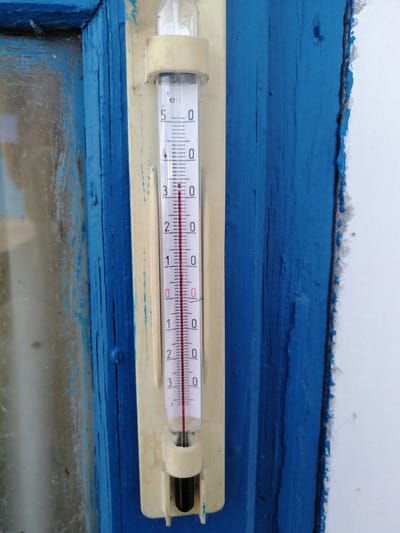 A vertical, glass thermometer with a red liquid column is mounted on a cream-colored plastic bracket. The bracket is attached to a weathered, bright blue wooden door frame. The thermometer scale ranges from -2 to 5 degrees Celsius, with markings every 0.2 degrees and labeled every 1 degree. The red liquid column indicates a temperature slightly above 3 degrees Celsius. The thermometer is positioned next to a window with a slightly hazy, greyish glass pane. The overall scene depicts an outdoor setting, likely on the exterior of a building in Zhabinka, Belarus, given the characteristic blue painted wooden structures often found in the region. The paint on the door frame is chipped and worn, suggesting exposure to the elements.
