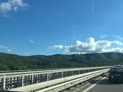 The image shows a view from a vehicle on a highway bridge, likely on the E81 road near Dolno Varshilo, Bulgaria. The bridge has metal railings. To the right, several cars are visible, including a black Mercedes-Benz with license plate C0 3446 BC. The landscape beyond the bridge consists of densely forested hills under a bright blue sky with scattered white clouds.