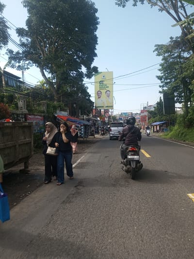 A road in Khe Luang, Laos, is observed during the day. Two women walk along the left side of the road, one wearing a hijab and the other covering her mouth with her hand. A motorcyclist travels down the right lane of the road. Several parked cars and shops are visible on the roadside. A political billboard is located above the buildings across the road. Trees line both sides of the road. The sky is clear.