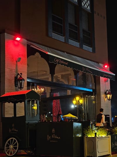 An eye-level outdoor shot depicts a restaurant at night. The building is light brown, with a dark green awning labeled "Cafe Restaurant" and "La Nome". A man in a vest walks past tables, some with green plants. In the foreground is a small cart with the "La Nome MARRAKECH" logo, and an ornate light fixture is mounted to the wall. Windows of the building have dark metal framing. Red lights are visible at the top and bottom of the frame.