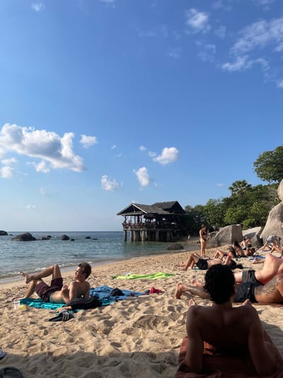 This image captures a bright, sunny beach scene in Tambon Ko Tao, Thailand. The foreground consists of golden-brown sand, where at least three individuals are prominently featured. In the immediate front, facing away from the camera, a person with dark, curly hair lies prone on a reddish-brown towel. To their left, a male with light brown hair lies on his back on a blue towel, wearing dark shorts, with legs bent and crossed; a plastic cup with a red straw, black sandals, and a black bag are beside him.

The midground expands to include a sandy beach populated by approximately 10-15 additional individuals, mostly lying on towels or directly on the sand, engaged in sunbathing. One woman in a dark bikini stands near the water's edge, facing towards a stilted building. The calm, blue-green ocean features several large, dark boulders. A prominent two-story wooden building with a dark, multi-layered shingled roof stands on stilts, extending partially over the water from the left side of the beach. Several people are visible on its open-air balconies. The background on the right is characterized by large, smooth, light-grey boulders and dense tropical foliage, including a palm tree. The sky is clear blue with scattered white cumulus clouds. Shadows on the sand indicate direct sunlight.