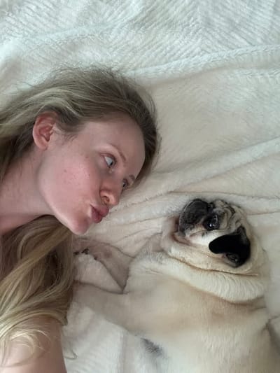 The media file is a close-up, overhead shot of a woman and a pug lying on a white, textured blanket. The woman is on the left side of the frame, her head tilted upwards, with her mouth puckered as if about to kiss or make a playful face. Her blonde hair is spread across the blanket. She has light skin with some freckles and is looking up and to the right. The pug is on the right side of the frame, facing the woman. Its fur is light-colored with darker markings around its eyes and ears. The pug's eyes are open, and it appears to be looking at the woman. The overall setting is intimate and casual, likely indoors, with the focus on the interaction between the woman and her pet. The lighting suggests daytime. No text is visible in the image.